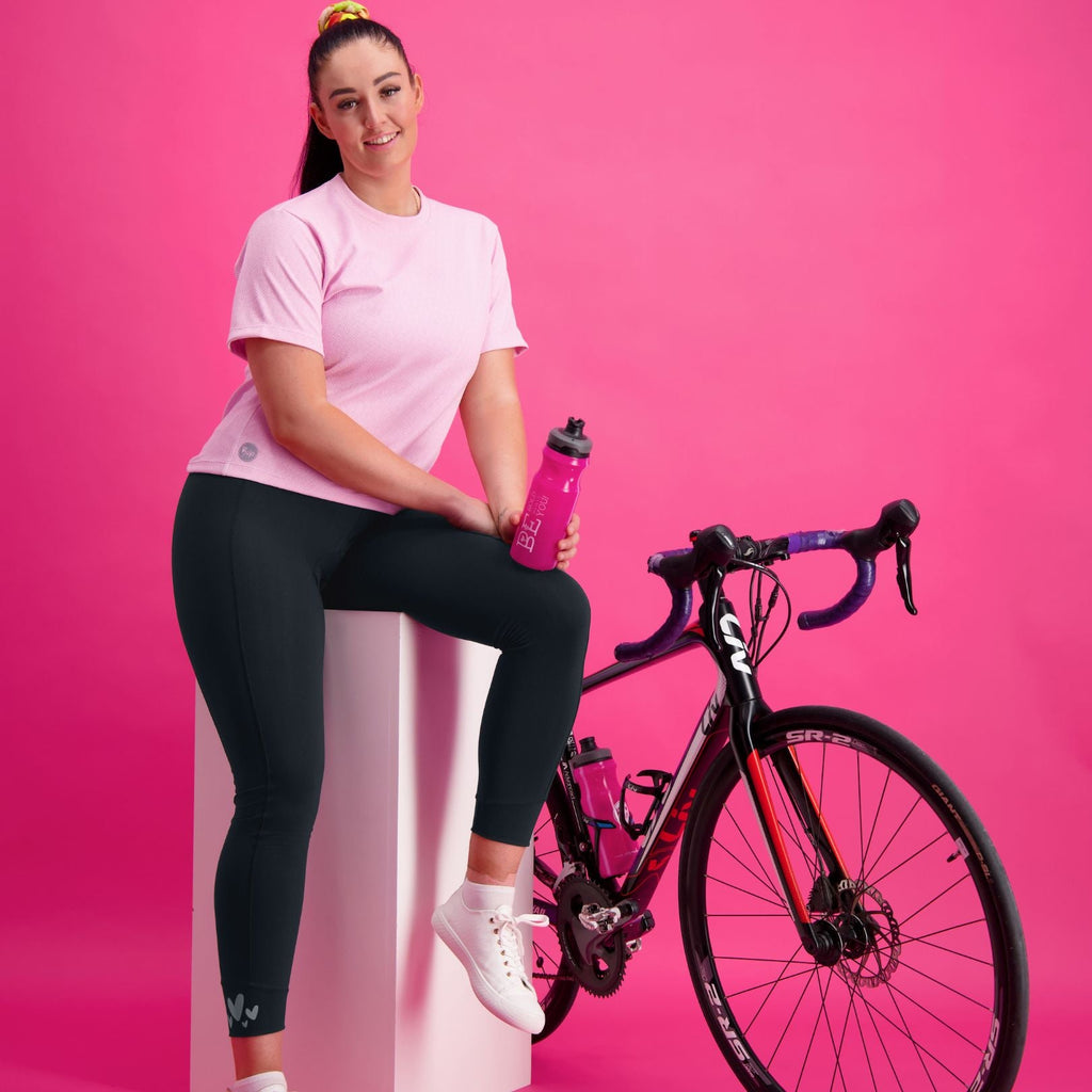 Woman with a pink top and charcoal grey cycling tights sitting on a box with a bike leaning on it